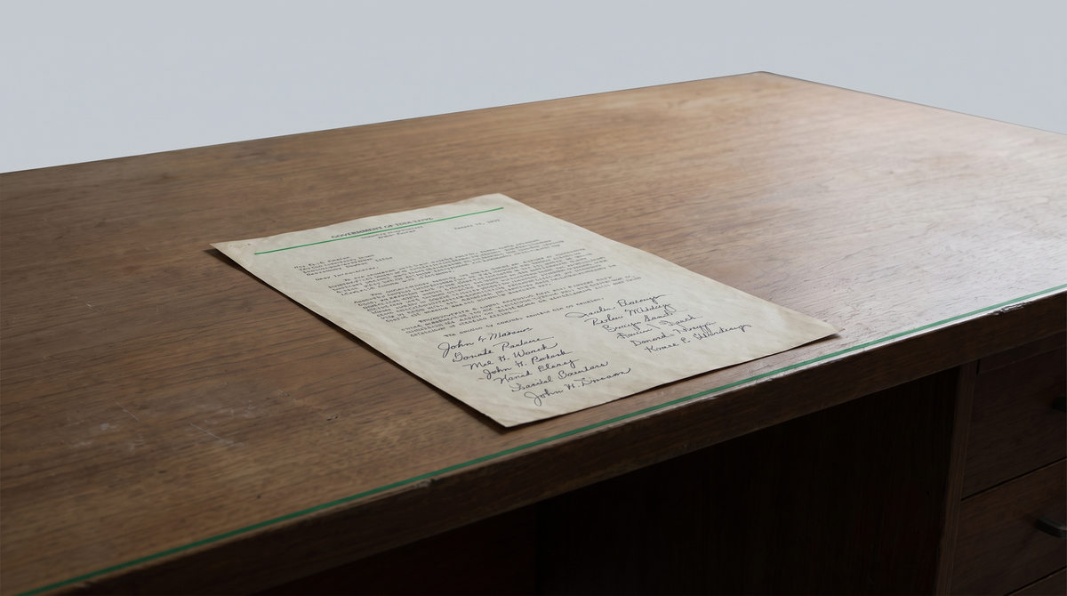 A formal typewritten letter with 16 signatures crossing a desk — on the other side an empty receiving surface — the space between the letter and the desk is the gap.