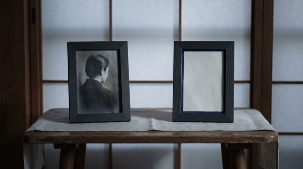 A photograph beside a blank space in a bedroom — the gap where a presence should be — a distance human grace cannot cross — deep indigo on grey